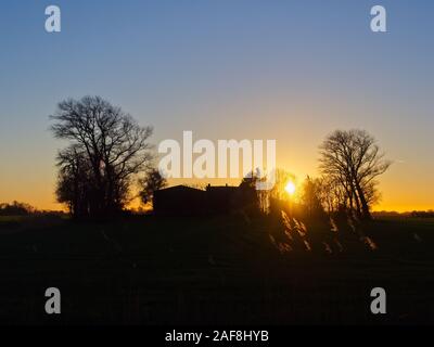 Silhoutte of a farm house between two trees with the sun setting behind it at Otterndorf Stock Photo