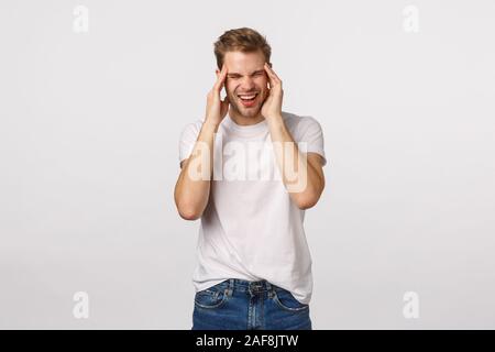 Man suffering headache after hangover, yesterdays party. Handsome blond man complain on migraine, squinting from pain, grimacing bothered, touch Stock Photo