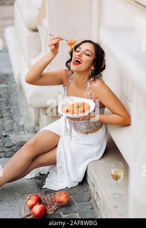 Beautiful smiling brunette girl having lunch in a street cafe, sitting at a table and eating Italian pasta. Advertising, commercial design. Stock Photo