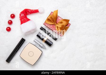 Christmas cosmetics background. Top view of various cosmetics products and beauty accessories, a gift box and a Santa hat on white snow. Space for des Stock Photo