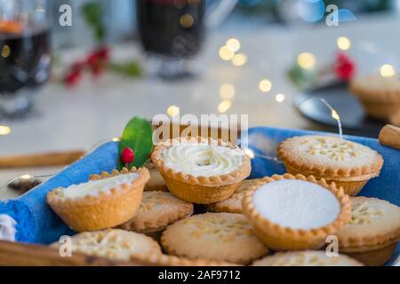 Close up traditional english festive pastry mince pies on wooden tray with blue napkin with blurred mulled wine drinks and lights garland on home tabl Stock Photo