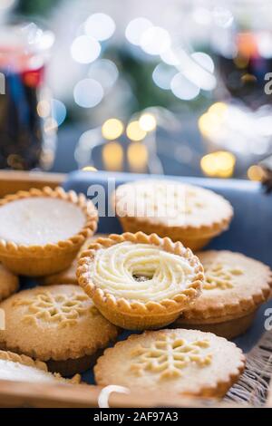 Warm cosy composition of traditional english festive pastry mince pies in wooden tray with blurred background of mulled wine drinks, lights garland an Stock Photo
