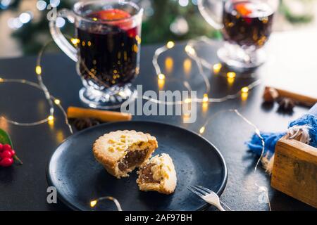 Close up broken Mince Pie with filling on black plate, mulled wine drinks and lights garland on dark table background. Traditional english festiveChri Stock Photo