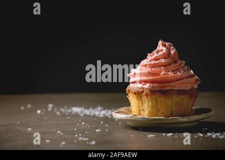 Homemade cupcake with pink buttercream and coconut flakes on ceramic plate on dark texture table. Copy space. Stock Photo