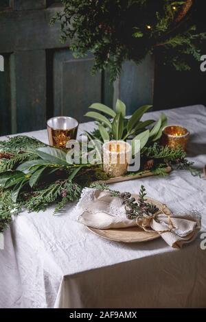 Christmas or New year table setting with empty ceramic plates, napkins, Christmas thuja wreath, luminous garland and burning candles on white tableclo Stock Photo