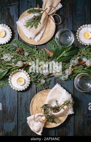 Christmas or New year table setting with empty ceramic plates, glasses, napkins, Christmas thuja wreath, luminous garland and burning candles on dark Stock Photo