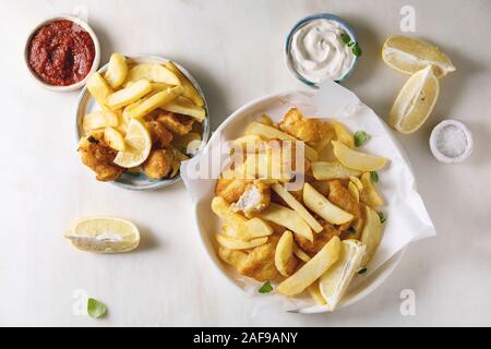 Classic british fast food fish and chips served on white paper in plate with lemons, red and white sauce, salt over white marble background. Flat lay, Stock Photo
