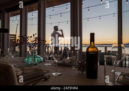 Dining table with wine bottle and glasses with man sipping red wine under deck lights at sunset in the background.