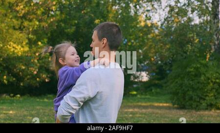 The little daughter is sitting in Dad's arms. She eats candy on a stick and gives to her dad. He doesn't want to, she laughs Stock Photo