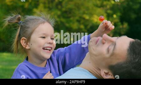 Close-up The little daughter is sitting in Dad's arms. She eats candy on a stick and gives to her dad. He doesn't want to, she laughs Stock Photo