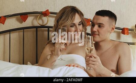 A young couple sits in bed, under a blanket. They're choking on champagne glasses and drink. Romantic date in a hotel room Stock Photo