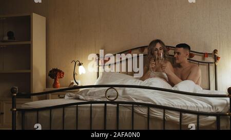 A young couple sits in bed, under a blanket. They're choking on champagne glasses. Romantic date in a hotel room Stock Photo