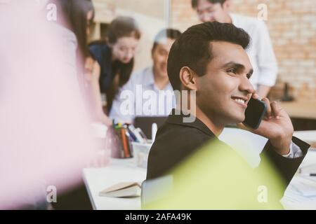 Indian businessman sitting in office and speaking on the phone with colleagues meeting in the background Stock Photo