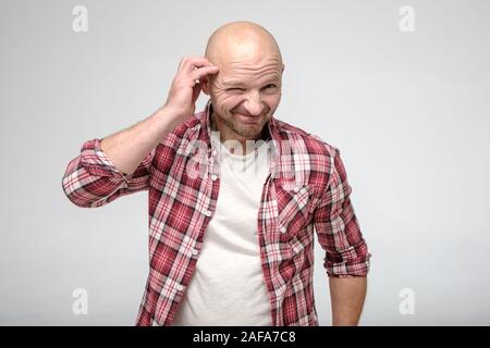 Bald man with stubble on face, looks into the camera with repentance, smiles guiltily and narrows one eye. Stock Photo