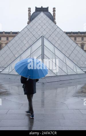 A man with a blue umbrella outside the Louvre and its famous glass pyramid entrance in Paris Stock Photo