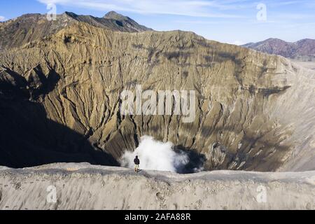 View from above, stunning aerial view of a person admiring the Mount Bromo with clouds of gases raising from the crater. Stock Photo