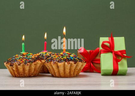Three tasty birthday cupcakes with chocolate glaze and caramel, decorated with burning festive candles and gift boxes on green background. Happy birth Stock Photo