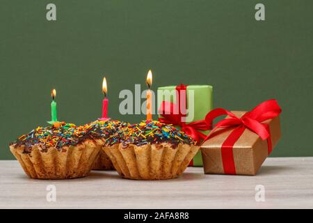 Three tasty birthday cupcakes with chocolate glaze and caramel, decorated with burning festive candles and gift boxes on green background. Happy birth Stock Photo