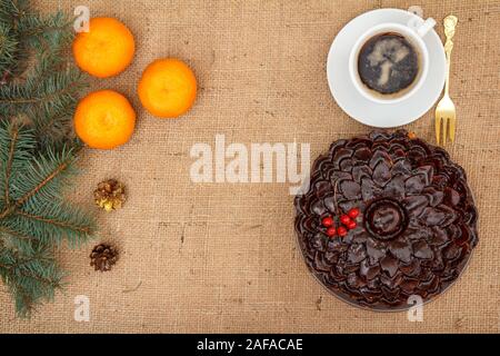 Chocolate cake decorated with bunch of viburnum, cup of coffee, oranges with branch of spruce on table with branch of spruce, fork and sackcloth.