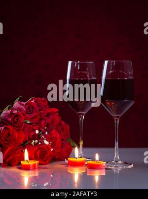 Bouquet of red roses and two glasses of wine with burning candles on a reflective white tabletop Stock Photo