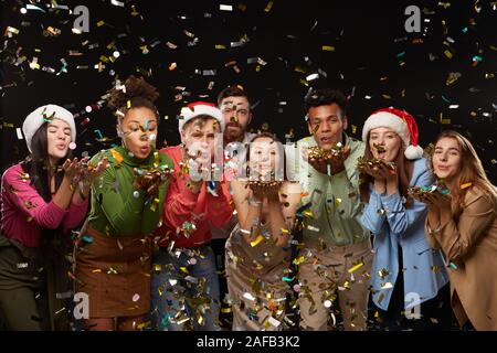 Group of cheerful young people are clinking with champagne glasses. On friends from above the festive confetti falling. Shooting in professional studio on isolated black background. Stock Photo