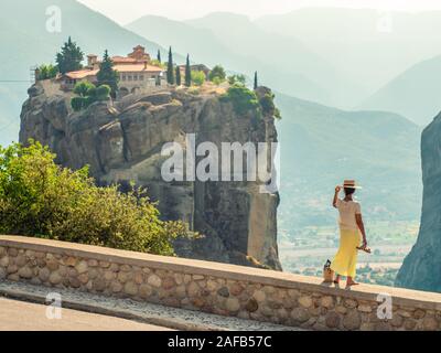 Young woman with white dress and large hat standing on wall in front of greece meteor mountains, monastery and village in the background Stock Photo