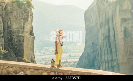 Young woman with white dress and large hat standing on wall in front of greece meteor mountains, monastery and village in the background
