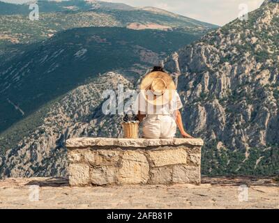Young woman in fashion dress sitting on stone becnch in front of Delfi mountain landscape in Delphi, Greece Stock Photo