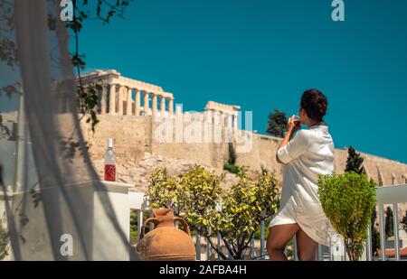 Young woman tourist having a drink on house balcony, with view of parthenon in Athens acropolis, Greece Stock Photo