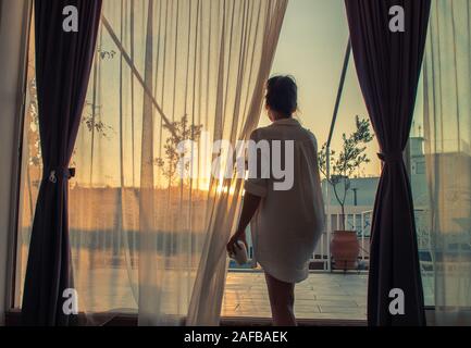 Young woman on a balcony looking at the sunset, view through transparent curtains Stock Photo