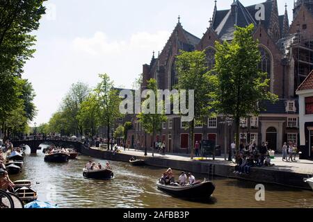 View of people riding a small boats on canal doing a cruise tour in front of 13th-century The Oude Church in Amsterdam. Many people hang out on street Stock Photo