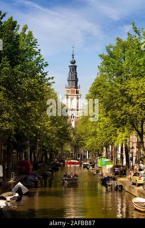 View of people riding a open boat on canal doing a cruise tour, Zuiderkerk church, trees and parked boats with blue sky background in Amsterdam. It is Stock Photo