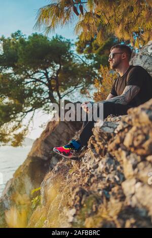 Handsome Tattooed Man Sitting On Cliff And Enjoying Sunset Over The Sea. Happiness and Meditation. Harmony With Yourself. Hiking Vacation. Stock Photo