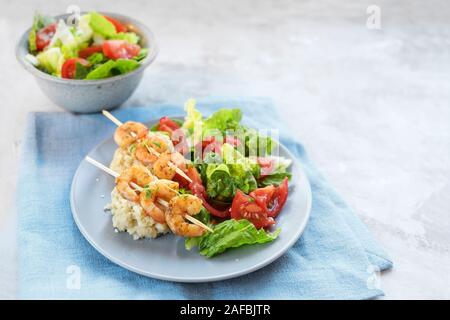 tiger prawn skewers on cauliflower risotto with salad of lettuce and tomatoes, plate and bowl on a blue napkin, light gray stone surface with copy spa Stock Photo
