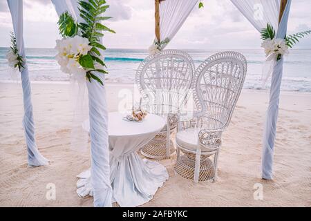 Decorated romantic wedding setting with table and chairs on sandy tropical beach with ocean and cloudy sky, Seychelles islands Stock Photo