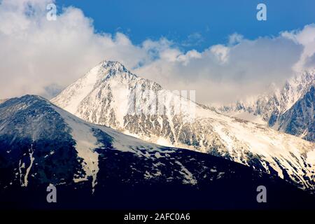 high tatras mountain ridge in springtime. snow capped rocky peaks in dramatic dappled sunlight beneath a clouds on a blue sky. place where earth meets Stock Photo