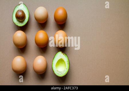 Raw two halves of avocado and brown chicken eggs on a brown background, flat, top view Stock Photo