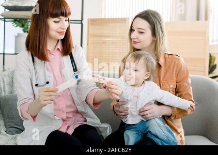 mother and baby toddler on reception at the child pediatrician. Female Pediatrician examining throat of a little ill girl. Kid looks sick and sad. Mot Stock Photo