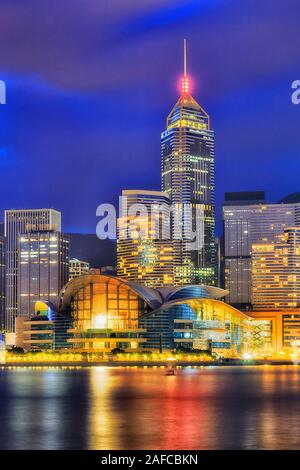 Bright illumination of modern urban buildings on Hong Kong waterfront - shores of Victoria harbour at sunrise against dark blue sky - vertical image w Stock Photo