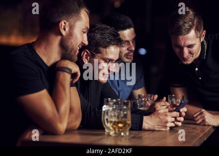 Funny content on smartphone. Three friends resting in the pub with beer in hands. Having conversation Stock Photo