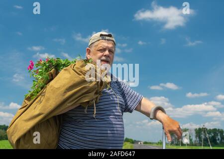 Portrait of Caucasian bearded senior man with sack and bunch of wild flowers standing on a roadside at summer season Stock Photo