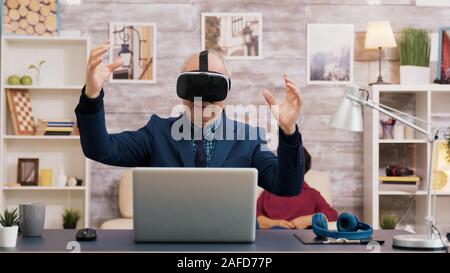 Senior man using virtual reality goggles in living room. Woman watching tv while sitting on the sofa in the background. Stock Photo