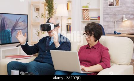 Elderly age woman sitting on sofa using laptop while her husband is experiencing virtual reality for the first time Stock Photo