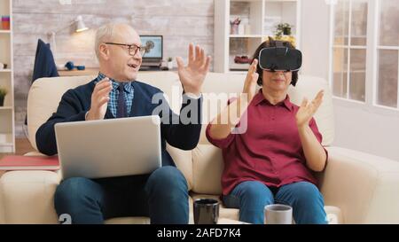 Amazed senior woman while using virtual reality goggles on sofa with her husband next to her using laptop. Stock Photo