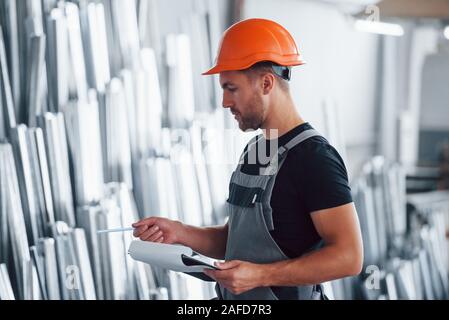 Counting and checking objects in the storage. Industrial worker indoors in factory. Young technician with orange hard hat Stock Photo
