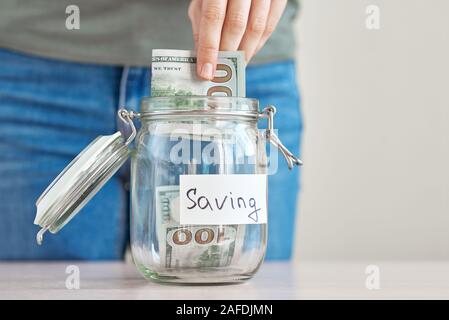 Woman hand holding us dollar bill and putting in glass jar with inscription saving. Saving money and home budget concept Stock Photo