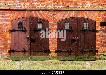 Close up image of a couple of wooden doors of a historical red brick building, with metal supports and locks, with moss on the wall and on the ground. Stock Photo