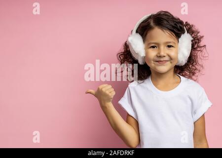 Positive little girl with curly hair, wearing white t shirt, furry earmuffs, pointing aside with thumb with cheerful expression, showing free space fo Stock Photo