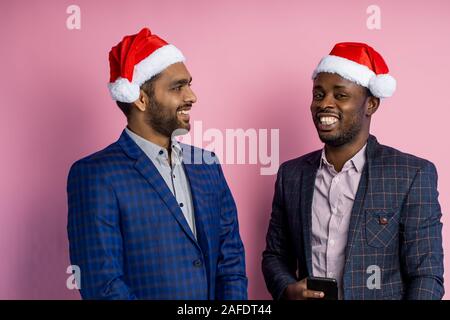 African american and indian man, wearing formal wear, standing against pink wall. Two colleagues in Santa hats, cheerfully communicating during break, Stock Photo