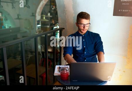 Young attractive hipster freelancer working remotely on laptop in loft cafe. Businessman browsing internet at coffeshop. Blogger posts to social media Stock Photo